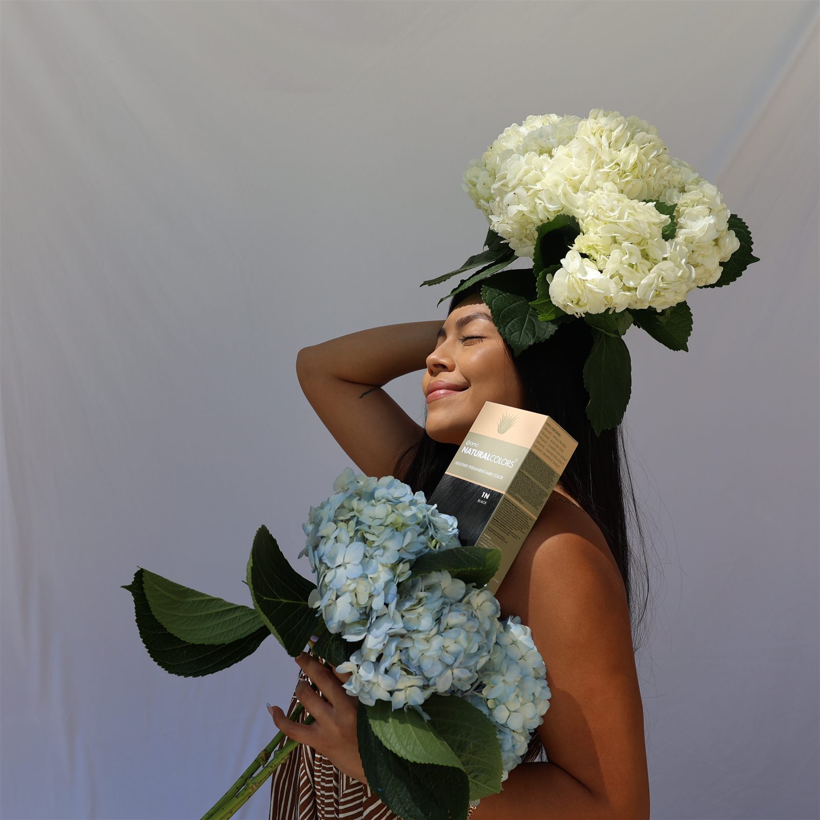 Model holding natural hair color product among blue and white hydrangeas